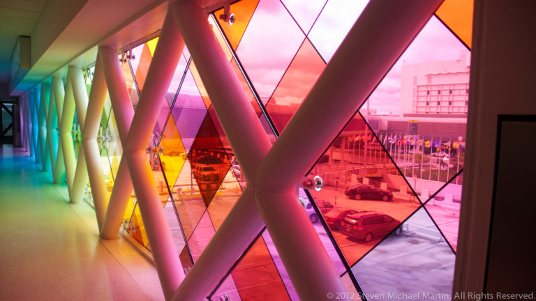 Airport corridor with multicolored geometric glass panels, diagonal supports, and aircraft visible outside.