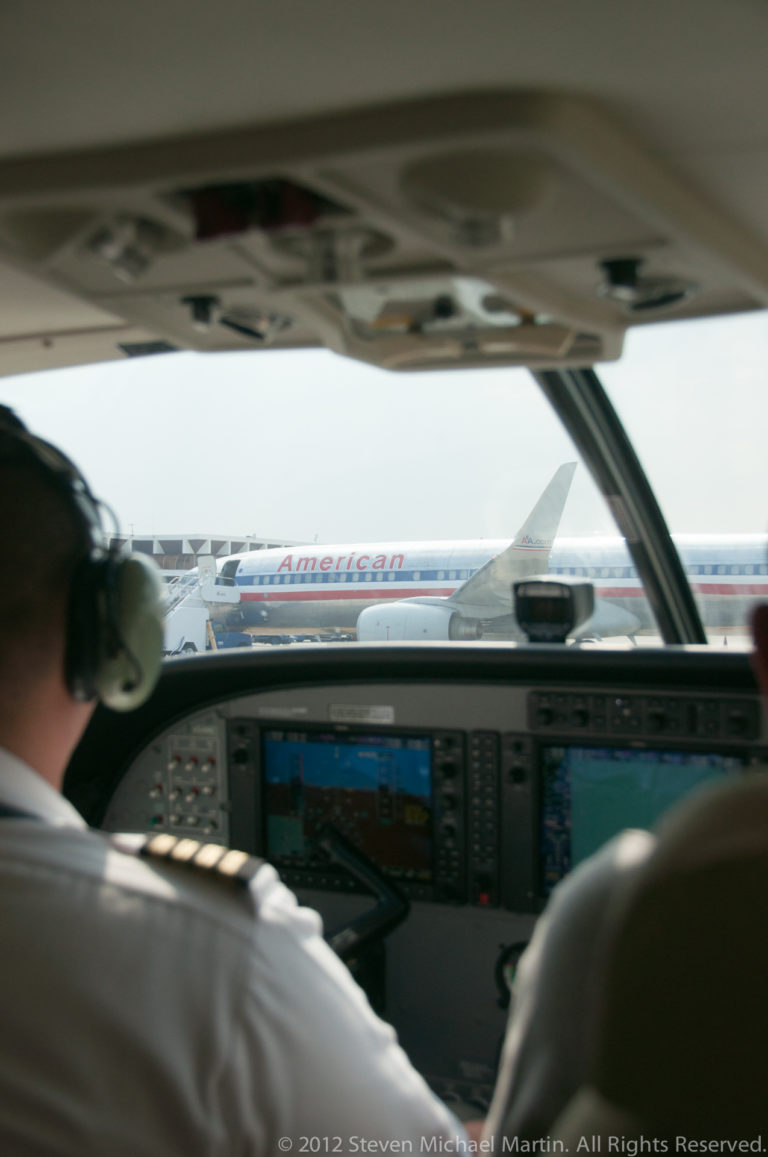 View past a pilot and cockpit instruments toward another aircraft parked outside on the apron.