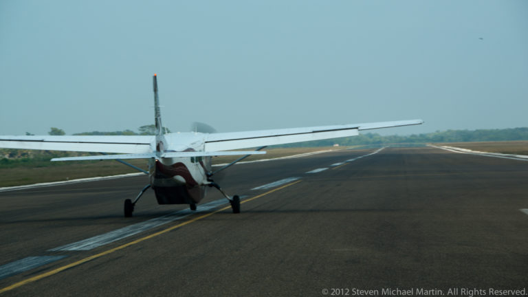 Small high-wing commuter aircraft viewed from behind on a paved Belizean runway.