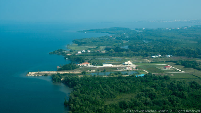 Developed Belizean shoreline with docks, buildings, dense vegetation, and deep blue coastal water.