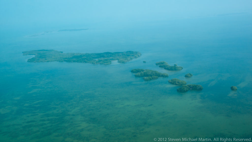 Cluster of small mangrove cayes scattered across pale turquoise Caribbean shallows.