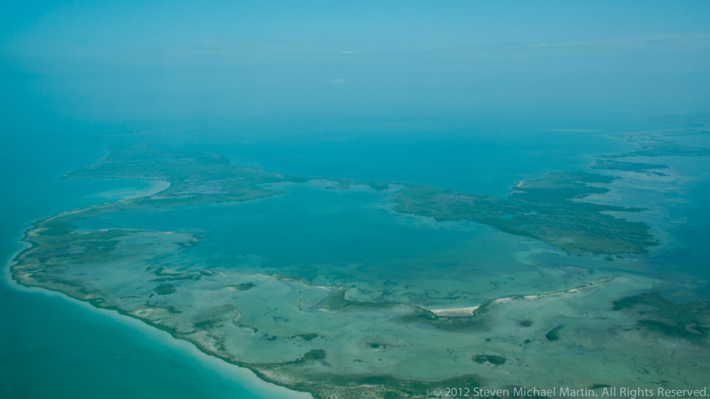 Low Belizean cayes and pale reef flats surrounded by turquoise Caribbean water in an aerial view.