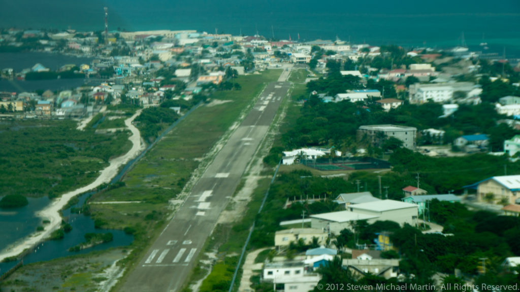San Pedro runway seen from an approaching aircraft, surrounded by roads, buildings, vegetation, and Caribbean water.