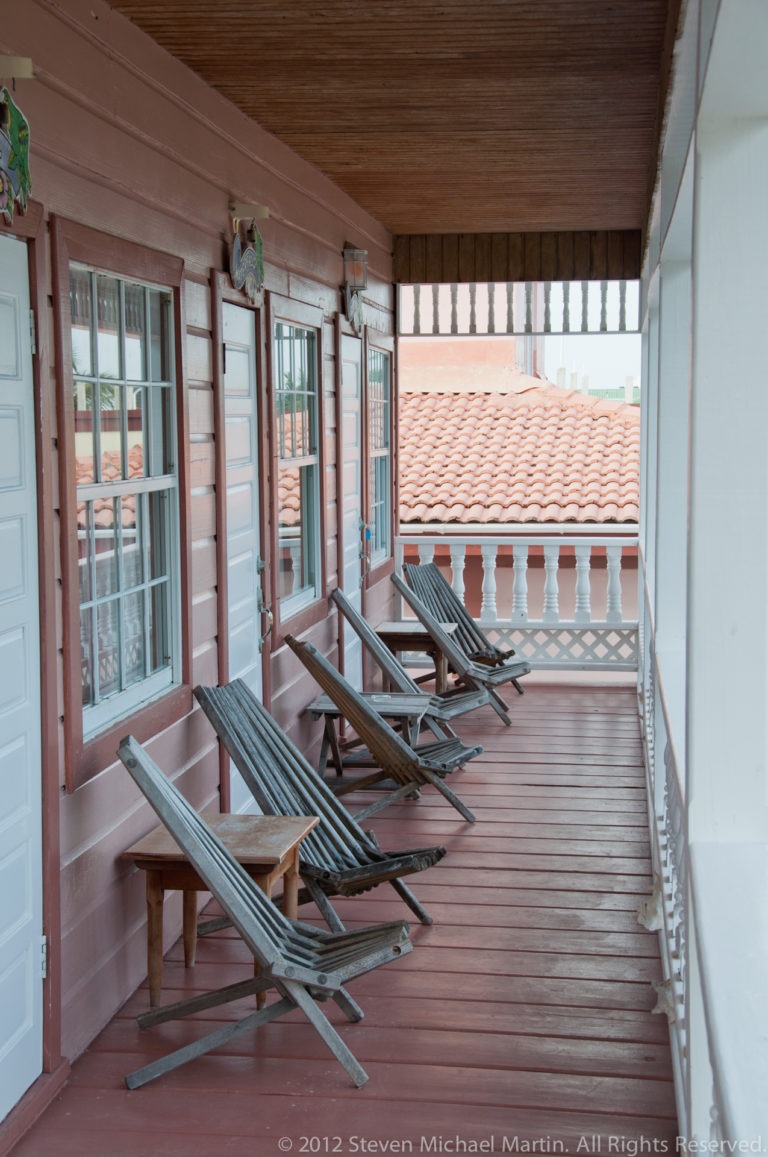 Wooden lounge chairs arranged along a shaded veranda with shuttered windows, turned railings, and plank flooring.
