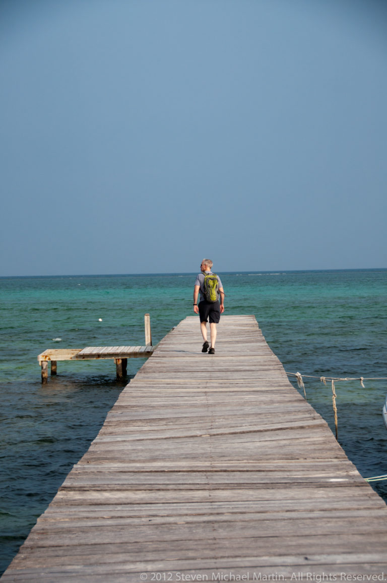Person crouching near the end of a long wooden pier surrounded by turquoise Caribbean water.