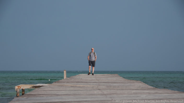 Solitary person standing near the end of a long wooden pier extending into turquoise Caribbean water.