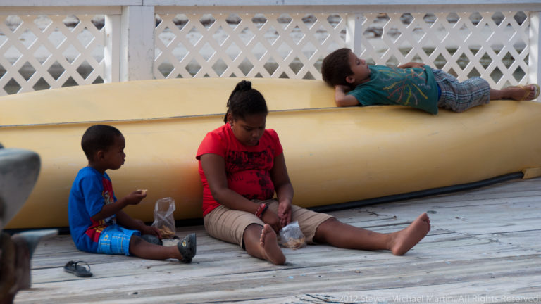Three children resting on a wooden deck beside a large yellow canoe in San Pedro.