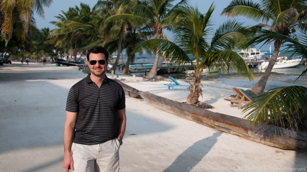 Man wearing sunglasses standing on a sandy palm-lined waterfront with boats and turquoise water behind him.