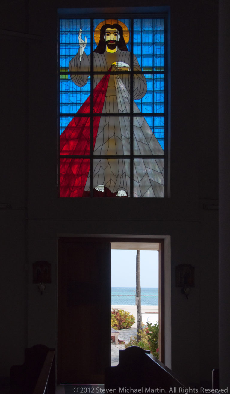 Stained-glass figure of Christ above an open doorway leading toward a palm-lined Caribbean shore.