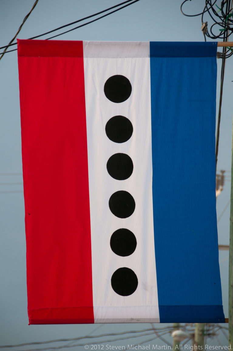 Red, white, and blue vertical banner with six black discs hanging beneath utility wires in San Pedro.