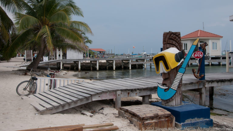Wooden waterfront footbridge beside a carved colorful sign, bicycle, palms, and piers in San Pedro.