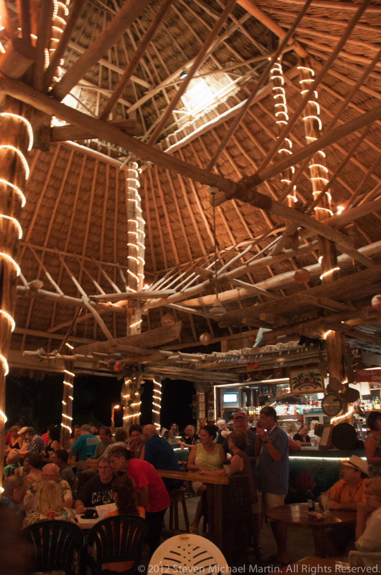 Busy open-air San Pedro restaurant beneath a high thatched roof with timber framing and string lights.