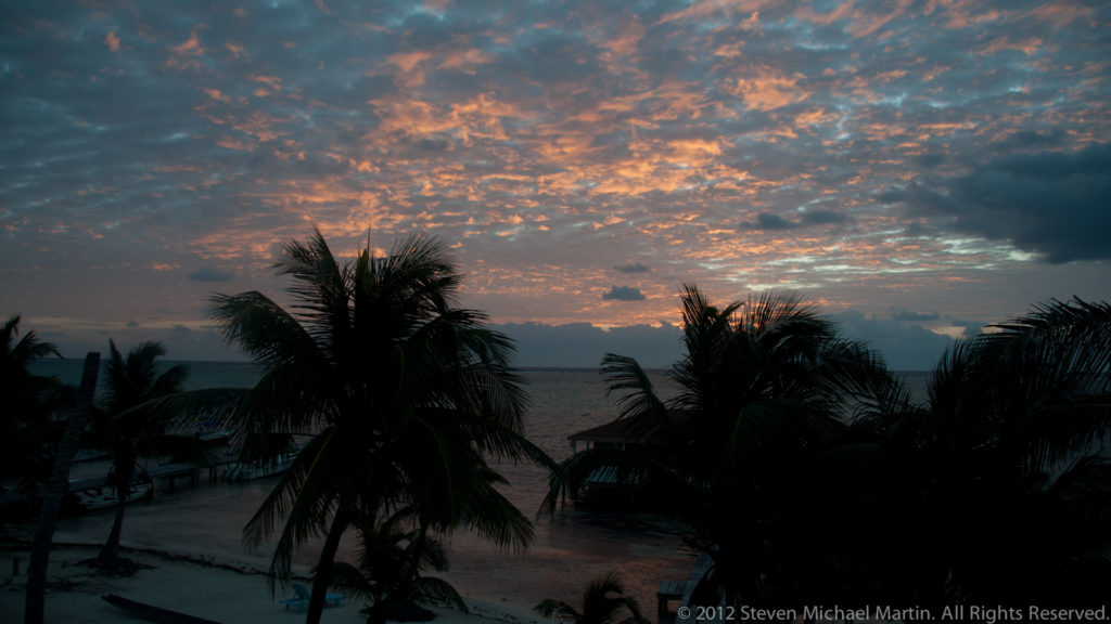 Palm trees and beach structures silhouetted beneath pink-orange dawn clouds on San Pedro’s shore.