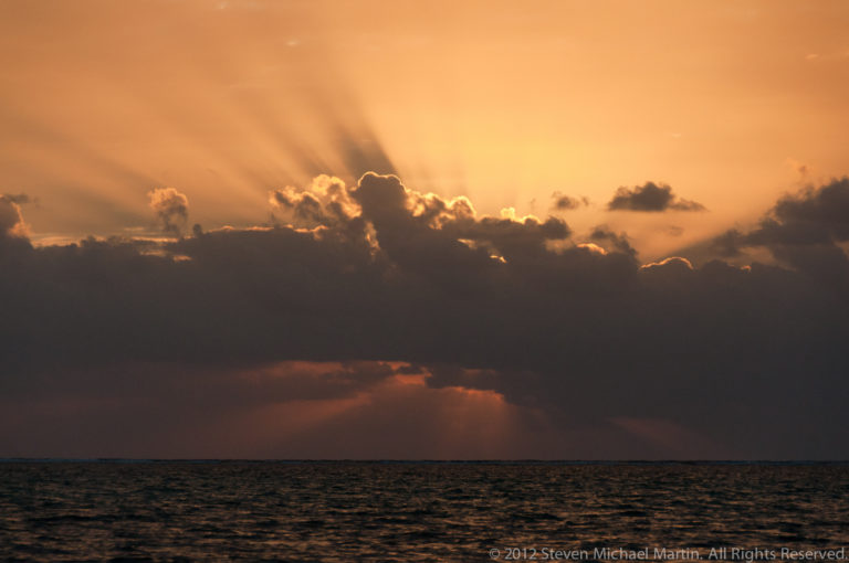 Broad golden sun rays fan behind dark clouds above a narrow strip of Caribbean Sea.