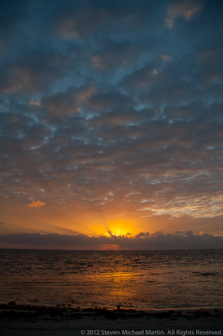 Orange sunrise band beneath layered clouds, reflected across rippled Caribbean water.