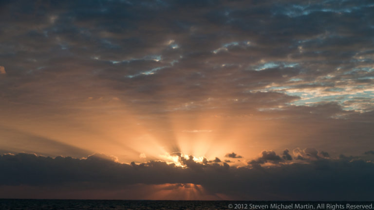 Golden sunbeams radiate through dark clouds above the Caribbean horizon at sunrise.