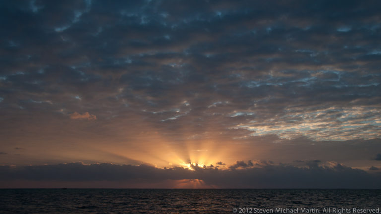 Sun rays fan upward behind low clouds over a dark Caribbean Sea horizon.