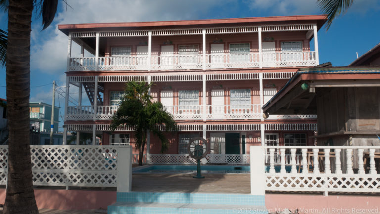 White multi-story San Pedro building with deep verandas, decorative railings, palm trees, and a red roof.