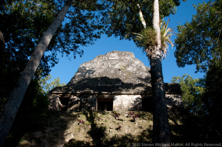 Small weathered limestone Maya temple at Tikal with a single central doorway, set on a low stepped platform beneath tall rainforest trees.