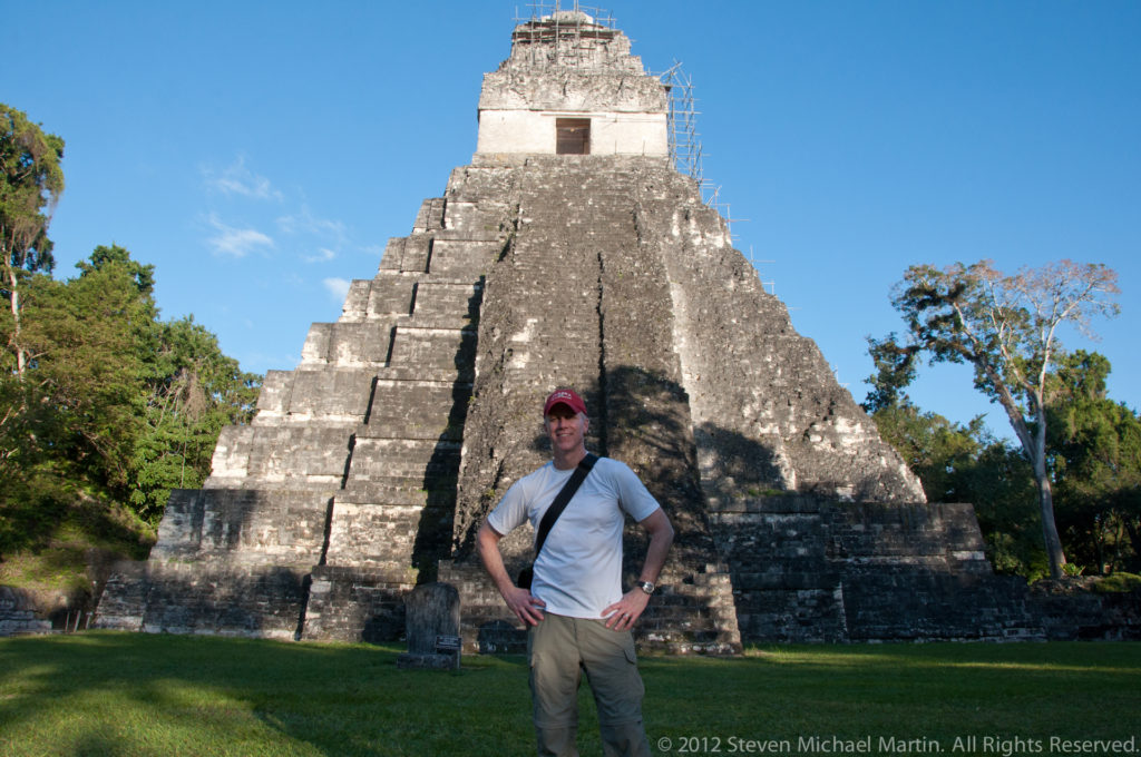Man standing on the lawn with hands on hips in front of a steep stepped Maya temple pyramid with a tall roof comb at Tikal, likely Temple II.