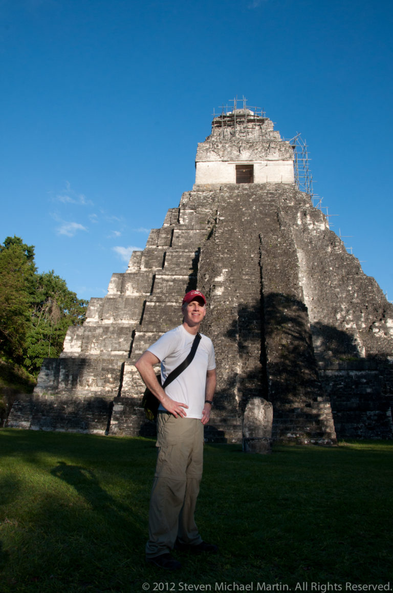 Man standing at the base of a steep Maya temple pyramid at Tikal, likely Temple II, beneath a clear blue sky.