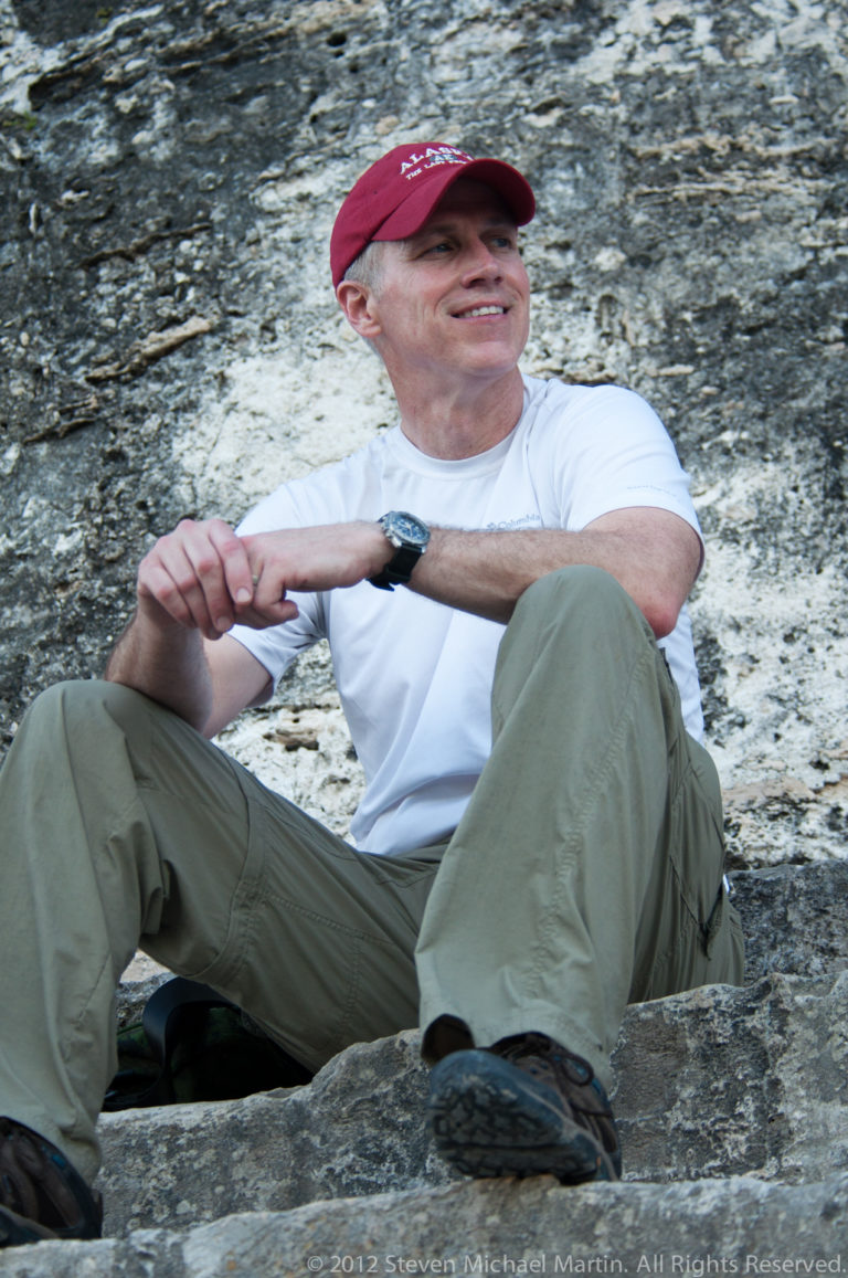 Cropped view of a man in a white shirt and green trousers sitting with clasped hands beside a weathered pale stone wall at a Maya ruin.