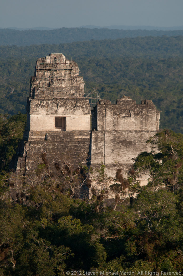 Telephoto view of a Tikal temple’s upper sanctuary and stepped roof comb rising above the surrounding rainforest canopy, with traces of pale stucco on the weathered masonry.