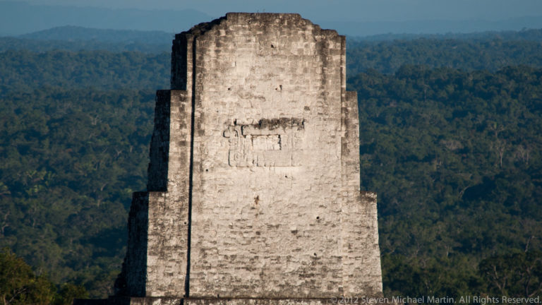 Telephoto view of a tall rectangular Maya roof comb of pale masonry towering above the forest at Tikal.