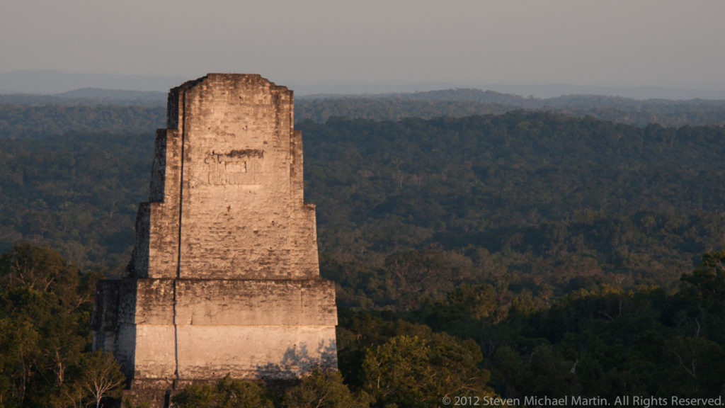 Distant view of a tall Maya temple roof comb at Tikal rising above dense rainforest canopy in warm light.