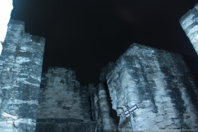 Artificially lit Maya stone walls at night, their rough limestone surfaces glowing against a black sky and forming a steep, enclosed space.