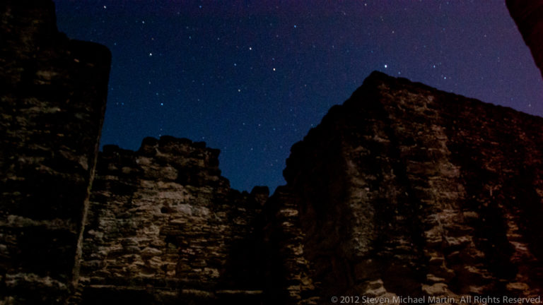 Night view of Tikal’s weathered Maya masonry walls in dark silhouette beneath a starry sky.