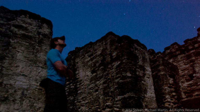 Man in a blue shirt looks up between tall, dark stone walls under a faintly starry night sky.