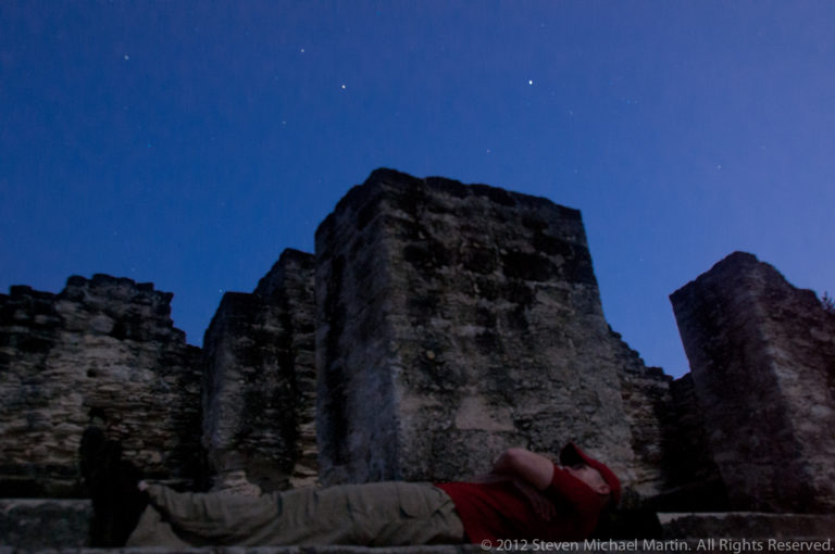 Visitor reclining beside low Maya stone ruins at Tikal under a dark blue, starry sky.