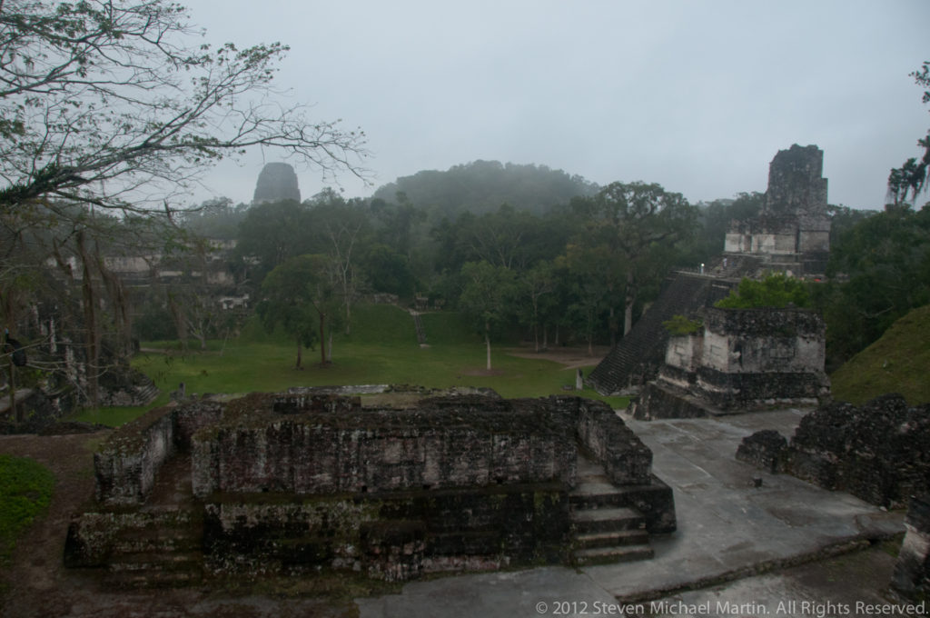 Elevated view over a broad Tikal plaza with low masonry platforms, open lawn, surrounding forest, and distant temples softened by mist.