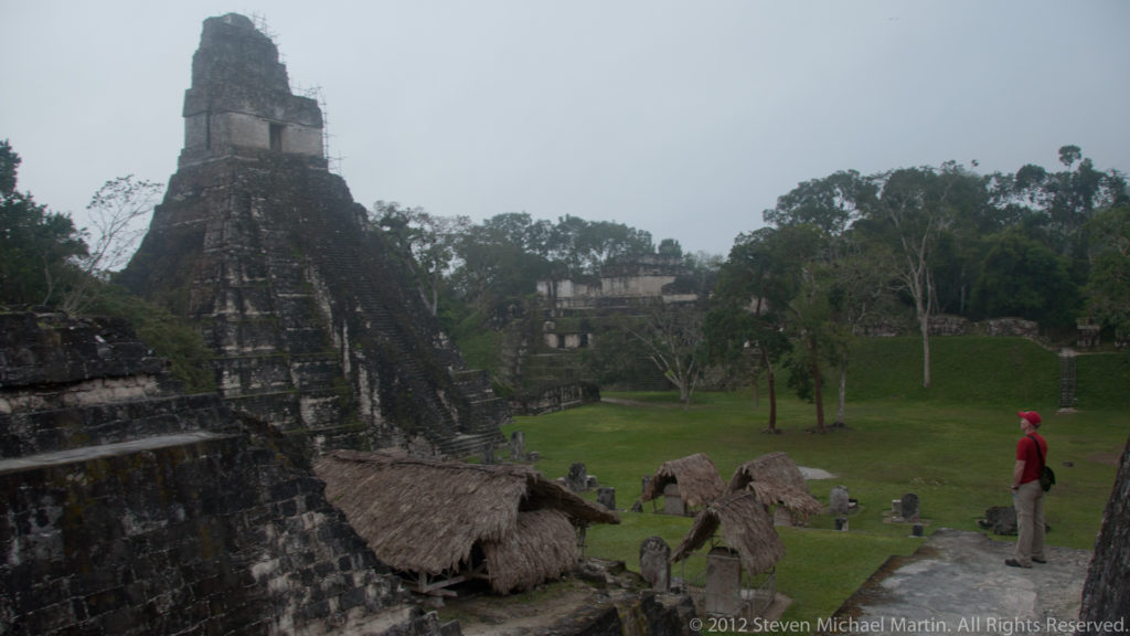 Elevated view over Tikal’s Great Plaza showing a tall temple pyramid to the left, an open grassy plaza in the center, and low masonry structures with thatched shelters in the foreground.