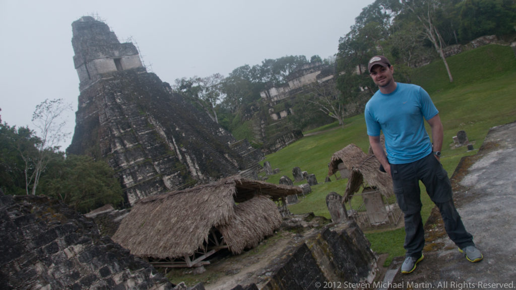 Visitor standing on an elevated ruin above Tikal’s Great Plaza, overlooking a tall temple pyramid, grassy plaza, and thatched shelters below.