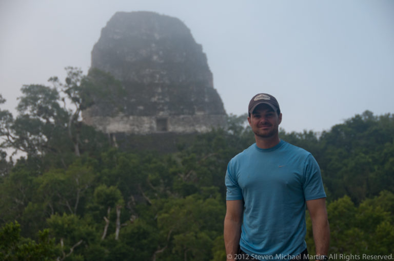 Man in a blue shirt standing facing the camera with a tall Maya temple pyramid rising into mist behind him at Tikal.