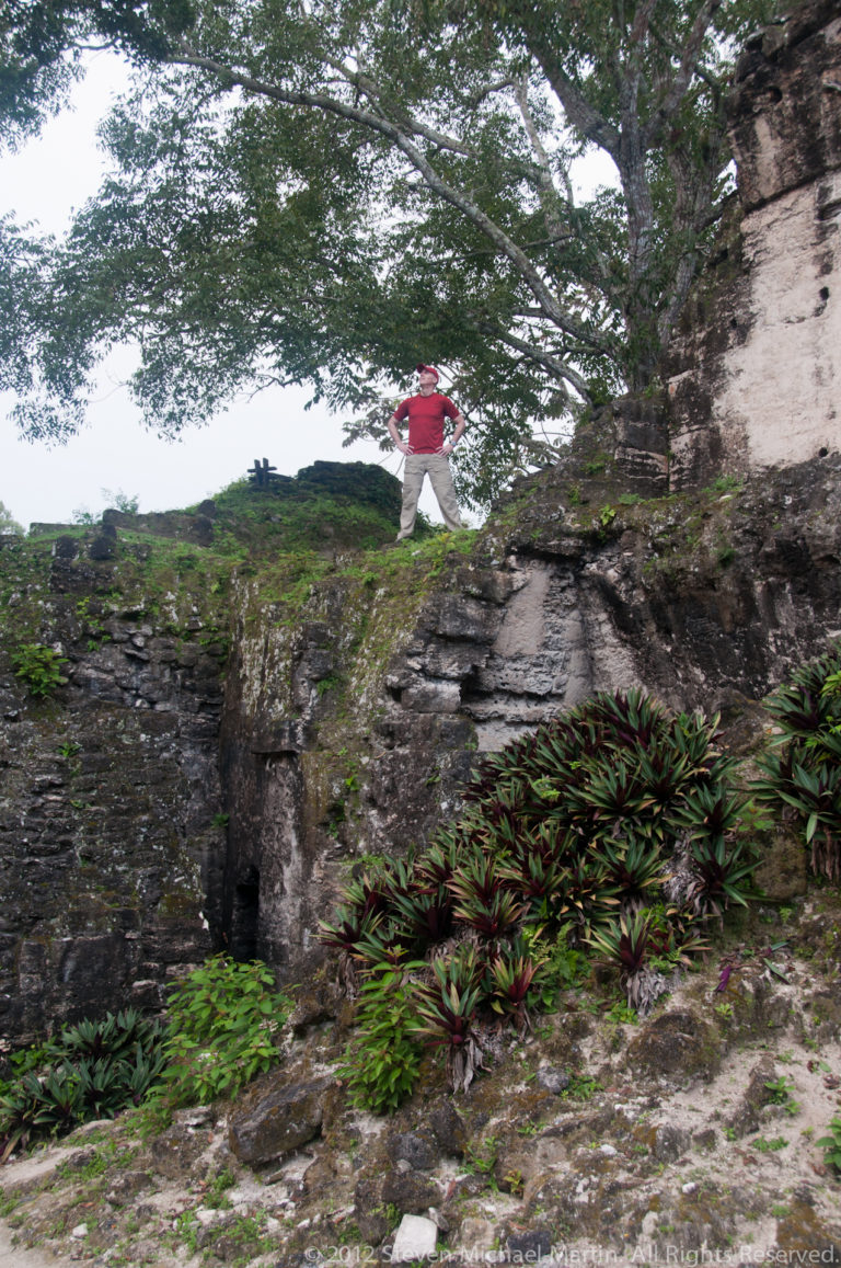 Visitor in a red shirt standing on the edge of a high, mossy stone platform above descending masonry terraces and ornamental vegetation at Tikal.