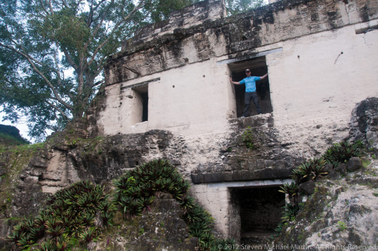 Visitor with arms outstretched in an upper doorway of a white-stuccoed Maya palace façade above a darker lower doorway.