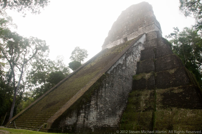 Low-angle view up a very steep stone staircase leading to the summit temple and roof comb of a Maya pyramid at Tikal in misty, wet conditions.