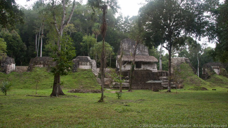 Low limestone Maya buildings and stairways surrounding a grassy courtyard in a forest clearing at Tikal.