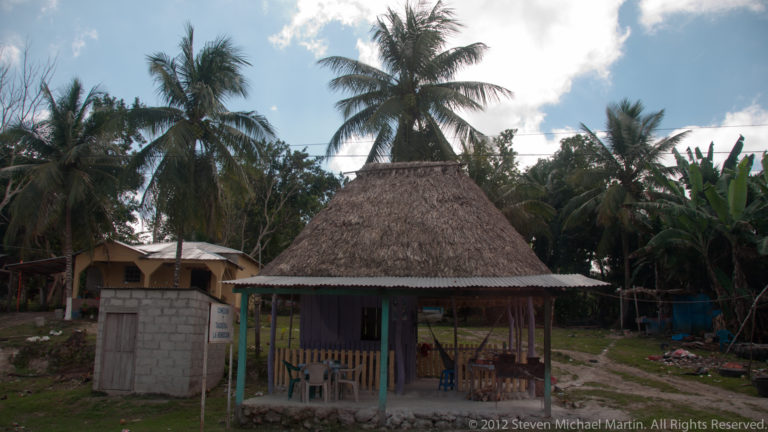 Small palm-thatched pavilion with chairs among coconut palms and simple buildings near Yaxha.