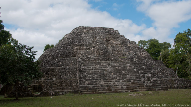 Broad stepped Maya pyramid with a central staircase, grassy foreground, and forest at Yaxha.