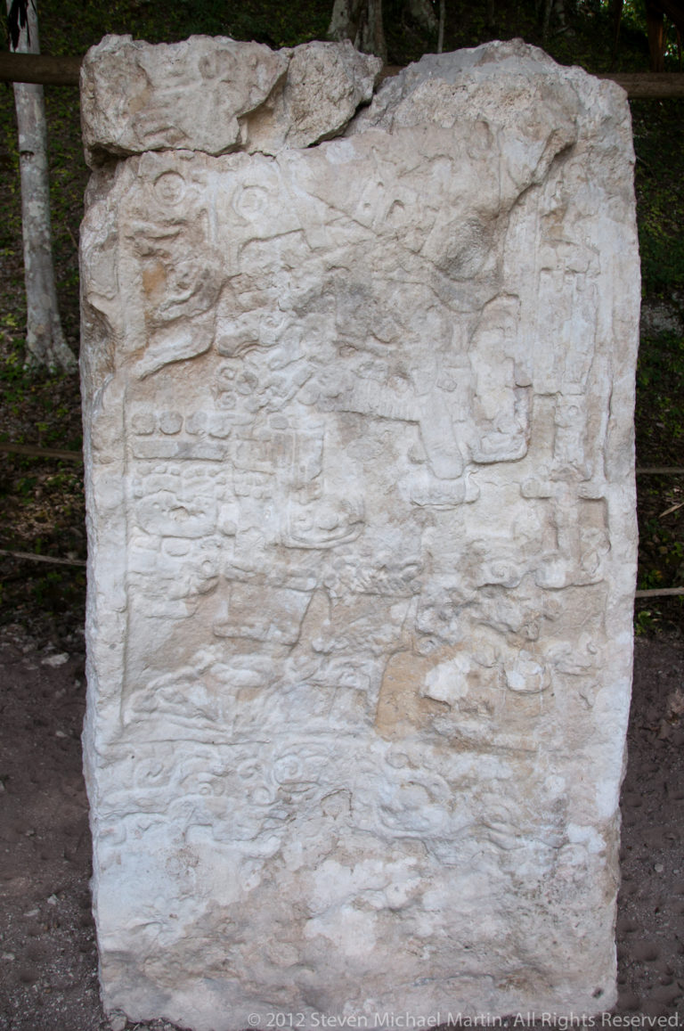 Weathered upright Maya stone monument with faint carved reliefs on a pale, eroded surface.