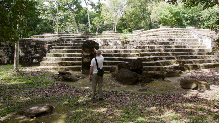 Visitor facing a low stepped Maya stone platform with large fallen blocks in the foreground.
