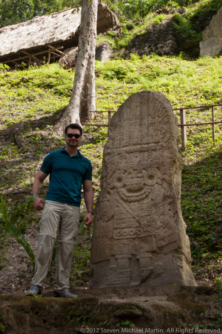 Visitor standing beside a tall weathered Maya stela with faint relief carving at Yaxha.
