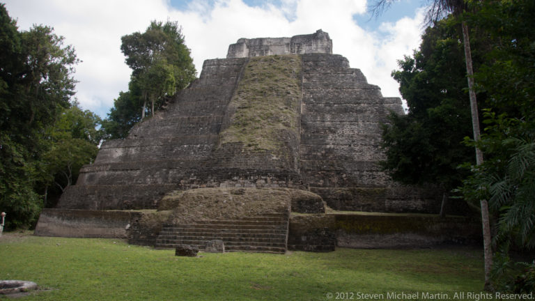 Tall steep Maya temple-pyramid with central stairs rising above a grassy clearing at Yaxha.