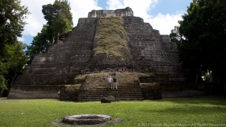 Two visitors at the base of a tall stepped Maya temple-pyramid with a central staircase at Yaxha.