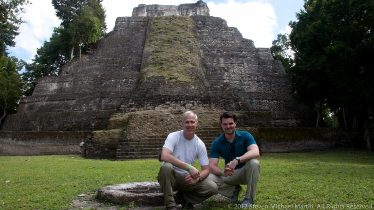 Two seated visitors in front of a towering stepped Maya temple-pyramid at Yaxha, Guatemala.