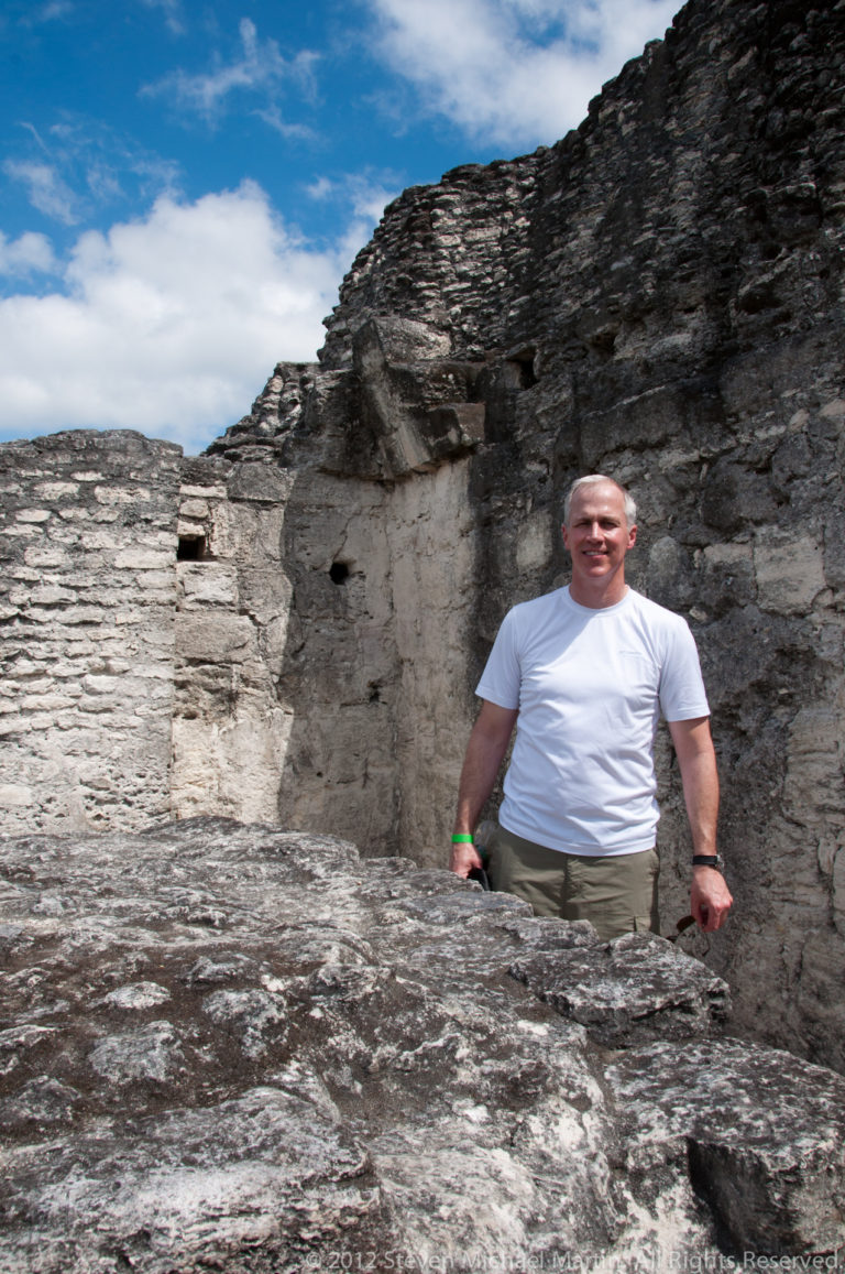 Visitor behind a low Maya stone wall with broken masonry and a rough opening at Yaxha.